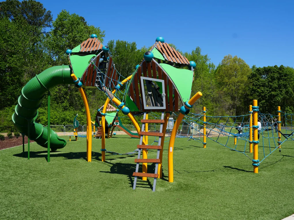 Modern Playground Equipment Colorful playground structure with a green tube slide, climbing nets, and platforms on artificial turf under a clear blue sky.