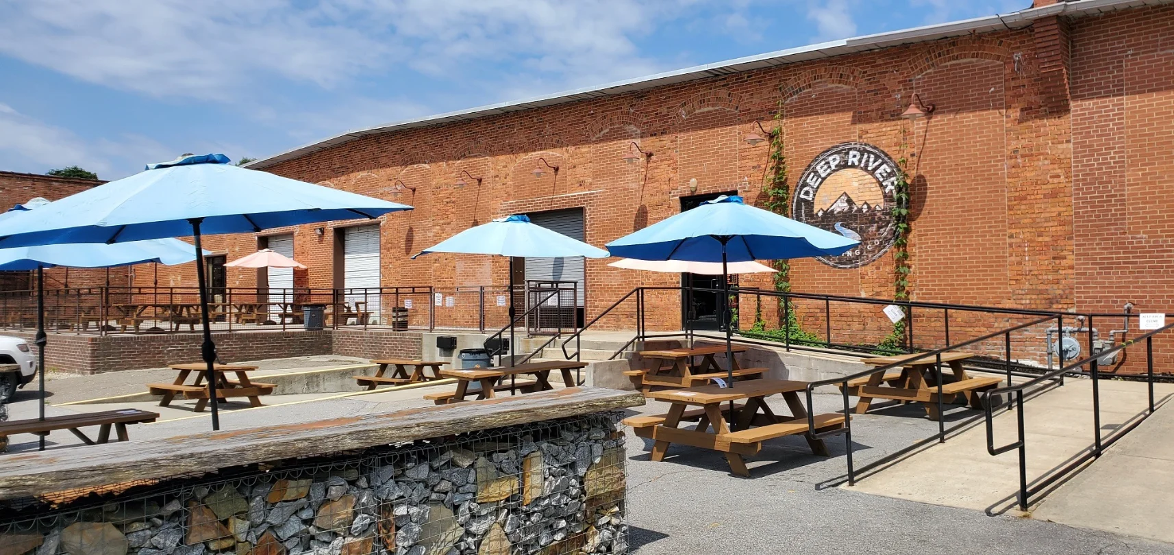 Deep River Brewing Co. Outdoor Seating Outdoor patio area of Deep River Brewing Co. with picnic tables, blue umbrellas, and a brick wall.