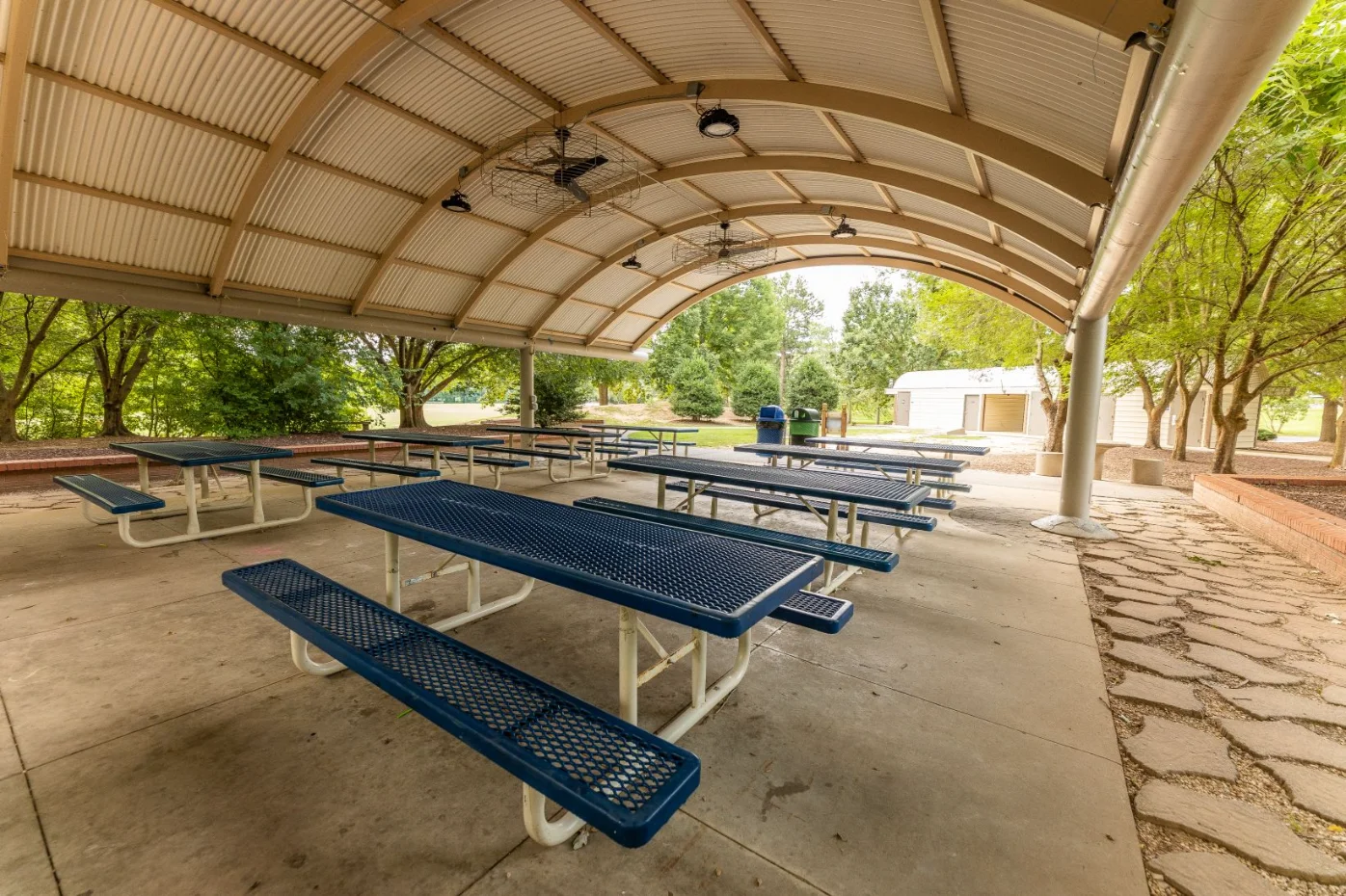 Community Picnic Shelter Covered outdoor picnic area with multiple blue metal picnic tables on a concrete slab under a large arched metal roof equipped with ceiling fans, surrounded by trees.