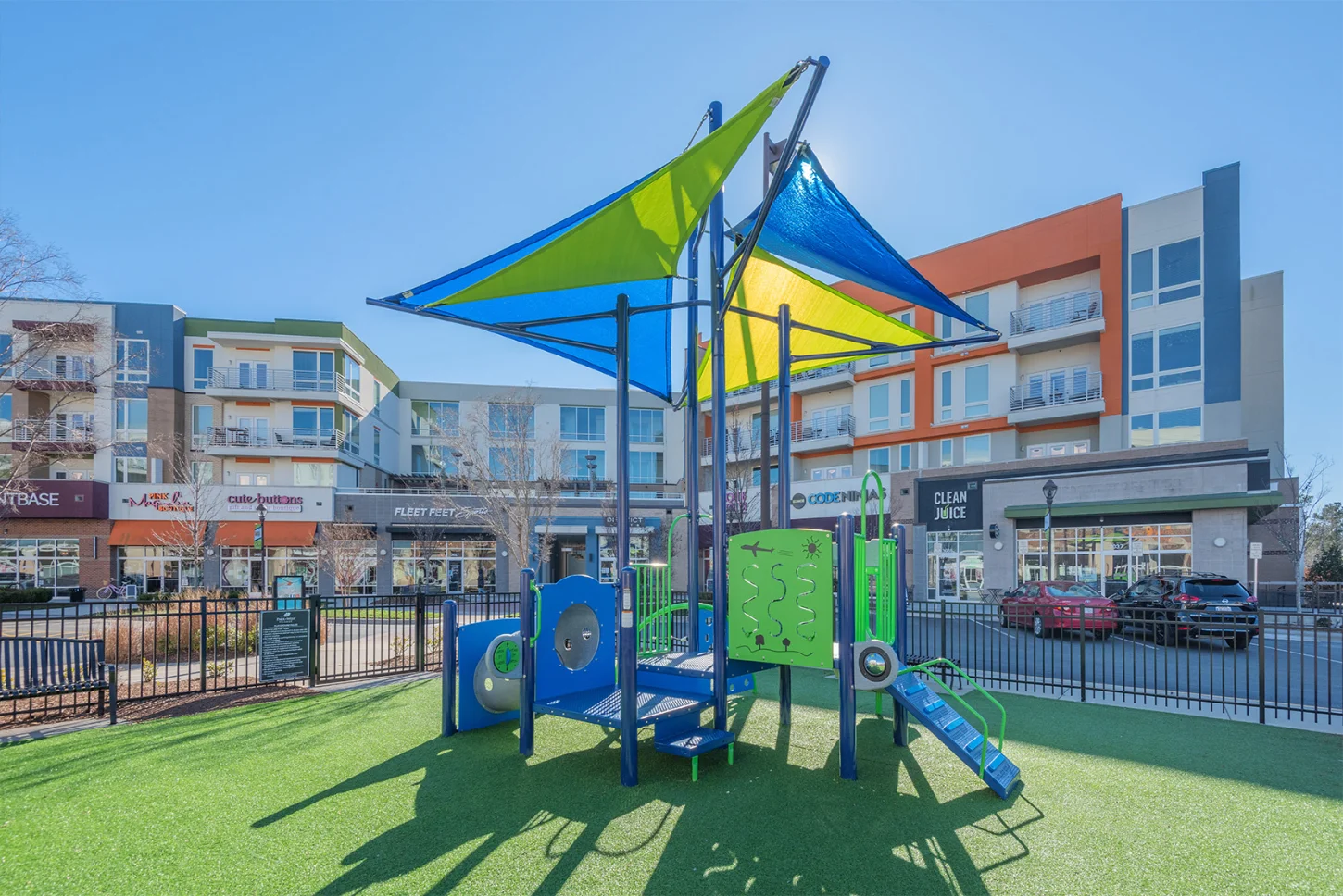 Urban Oasis: Modern Playground Colorful children's playground with blue and green equipment and yellow and blue shade sails, set on artificial turf, with modern multi-story residential and commercial buildings in the background on a sunny day.