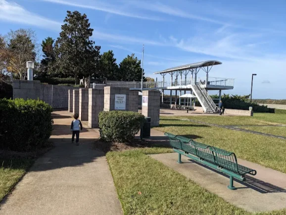 Park Exploration and Viewing Platform Young child walks on a paved path in a park towards a series of stone pillars and a modern elevated metal viewing platform in the background; a green park bench is in the foreground and a 'Smoker Facility' sign is visible.