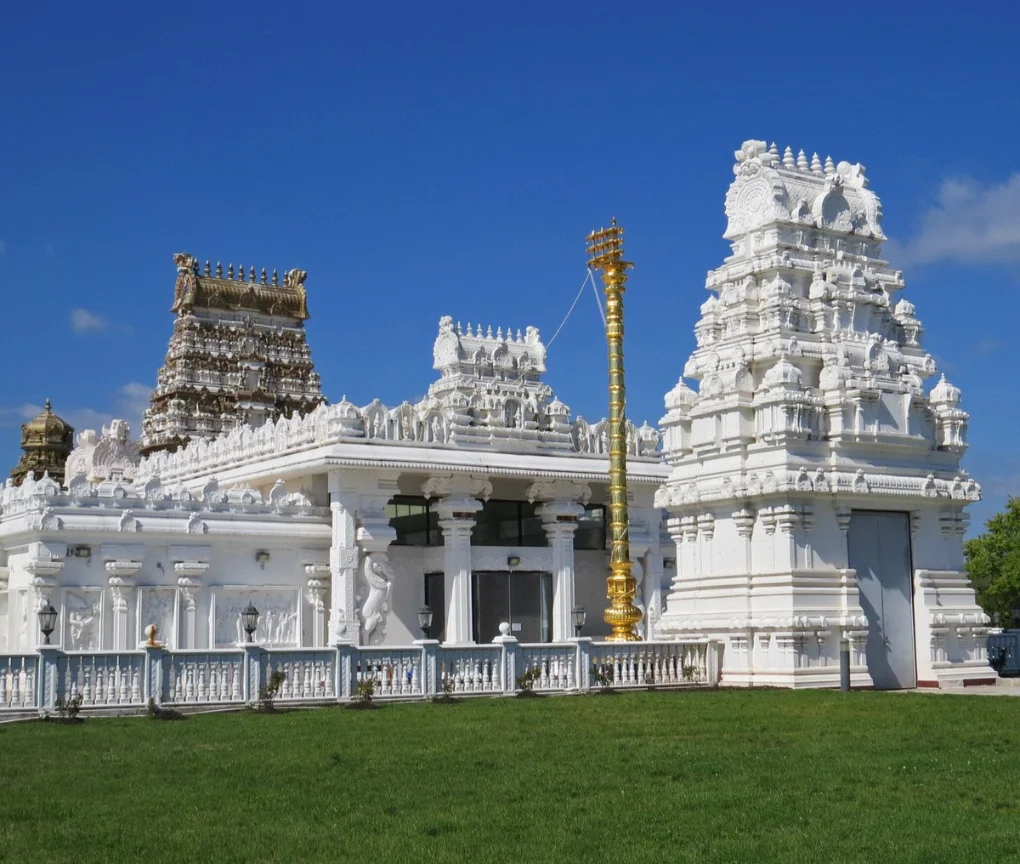 Sri Venkateswara Temple Magnificent white Hindu temple with intricately carved gopurams (towers) and golden embellishments, set against a clear blue sky with a green lawn in the foreground." Title: "Ornate Hindu Temple Architecture
